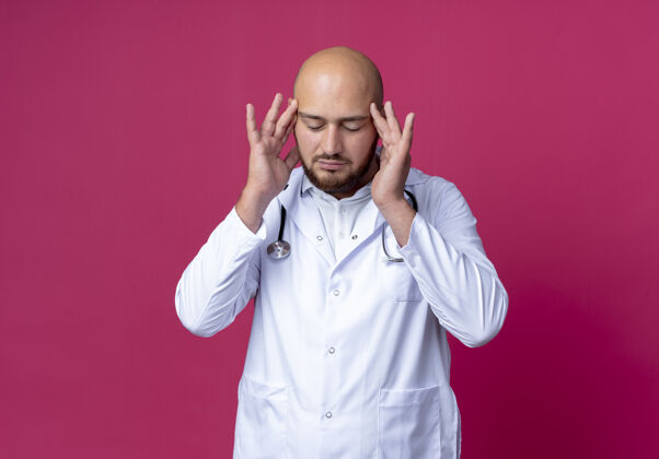 长袍年轻的秃头男医生戴着医用长袍 戴着听诊器 把手指放在额头上 隔离在粉红色的地方 留着复印空间复制疲倦粉色