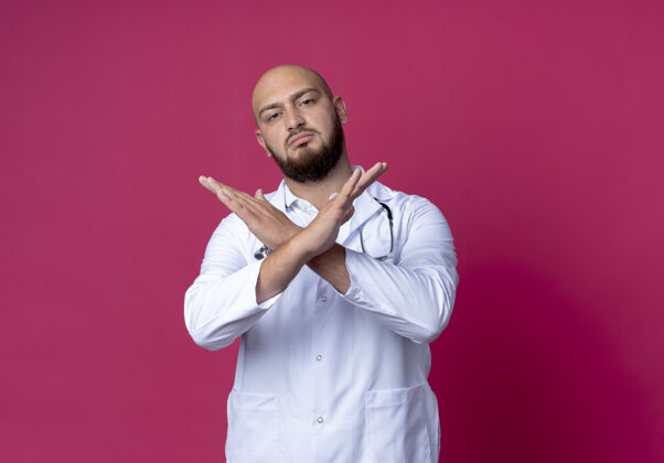 听诊器年轻的秃头男医生穿着医用长袍和听诊器 在粉色上显示出不孤立的姿态秀秃头粉色