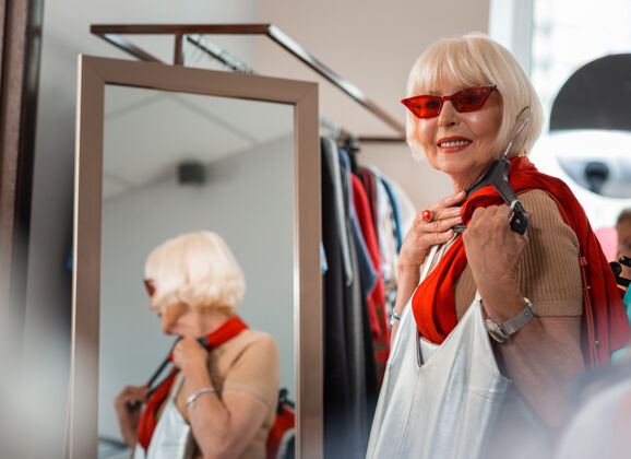商品爱上购物高兴的老妇人腰向上透过红色太阳镜看着别处 同时把新衣服放在胸前年龄成熟购物