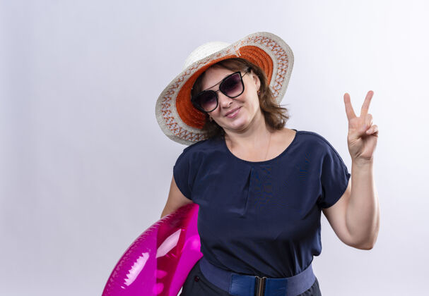 穿一位戴着眼镜 戴着帽子 手持充气戒指的中年旅行家女士在与世隔绝的白墙上展示着和平的姿态和平戒指展示