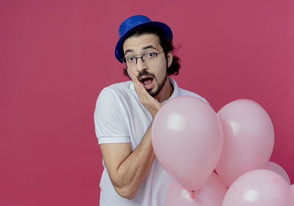帅气惊讶的帅哥戴着眼镜 戴着蓝色的帽子 手里拿着气球 把手放在下巴上 隔离在粉色上男人下巴戴着