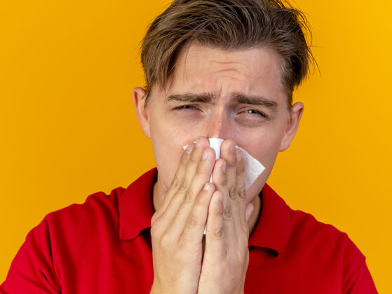 英俊特写镜头：悲伤的年轻英俊的金发病患 用隔离在橙色墙上的餐巾擦鼻子擦布疾病鼻子