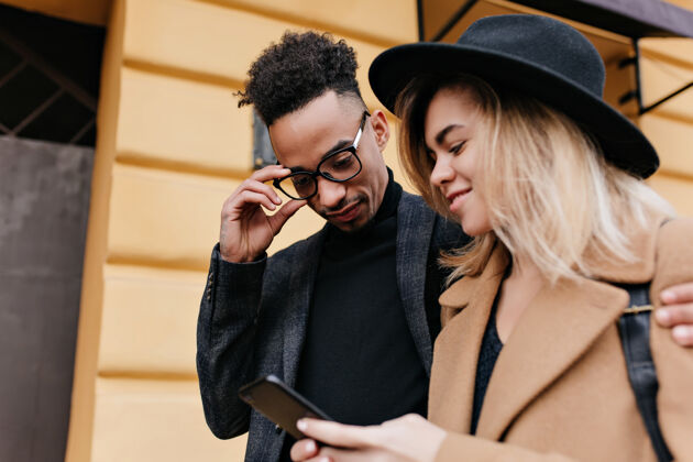 朋友快乐的白人女孩为非洲男性朋友展示新手机的户外写真时髦的戴眼镜的黑人年轻人在城市街道上和金发女郎玩得很开心休闲非洲金发