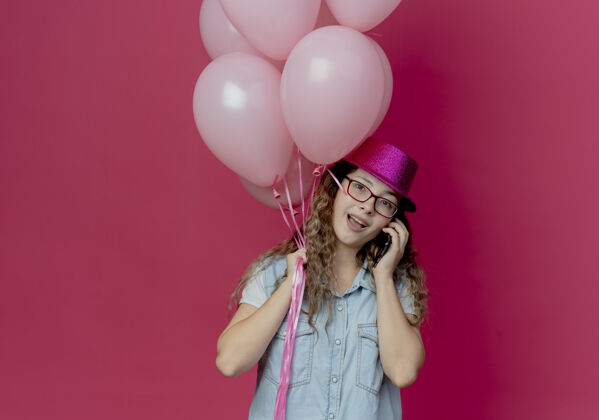 微笑微笑的年轻女孩戴着眼镜 戴着粉红色的帽子 手持气球 在电话里说孤立的粉红色年轻粉色帽子
