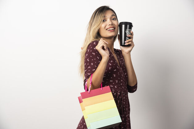 杯年轻女子拿着一个小购物袋和一杯饮料在白墙上购物者女人人