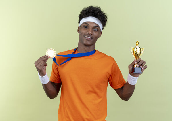 杯子快乐的年轻美国黑人运动男子戴着头带和腕带和奖牌杯隔离在绿色的墙壁上头带墙运动