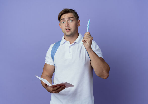 印象印象深刻的年轻帅哥学生背着书包拿着笔记本举着笔孤零零地站在蓝色的墙上笔墙姿态