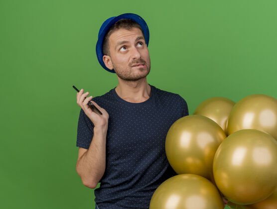 脸一个戴着蓝色派对帽 手持氦气球和手机 孤零零地看着绿色墙壁的困惑帅哥帽子氦电话
