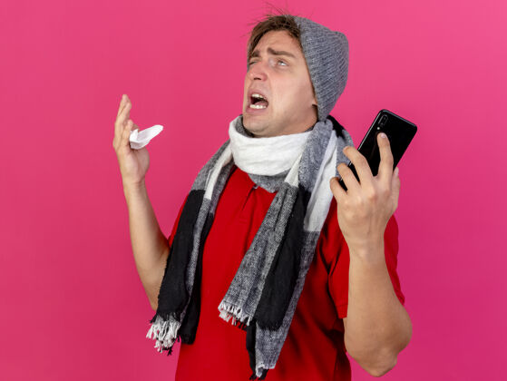 感情愤怒的年轻英俊的金发病患戴着冬天的帽子和围巾拿着手机和餐巾孤立地看着粉红色的墙上帽子围巾年轻