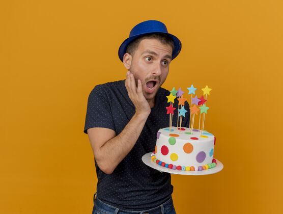 聚会焦急的帅哥戴着蓝色的派对帽 拿着生日蛋糕 看着橙色墙上孤立的一面举行帅气感情