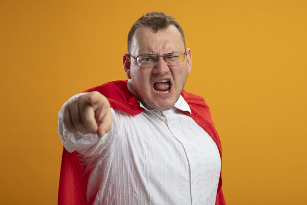 脸愤怒的成年超级英雄 穿着红色斗篷 戴着眼镜 指着橙色墙上孤立的前方愤怒人人