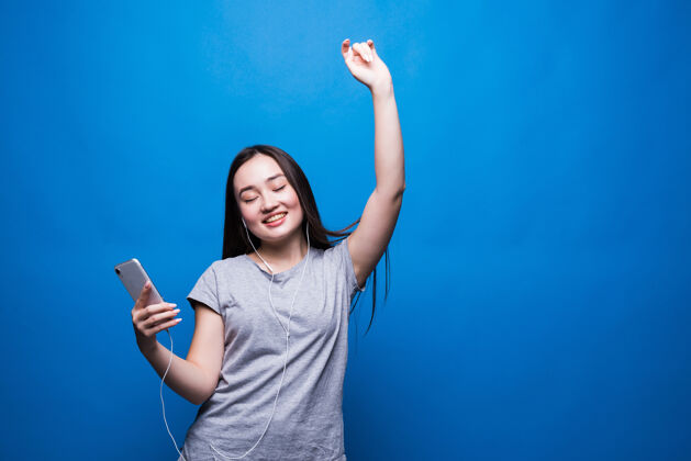 积极快乐的亚洲年轻女子戴着耳机听音乐 隔着蓝色的墙跳舞休闲全面音乐
