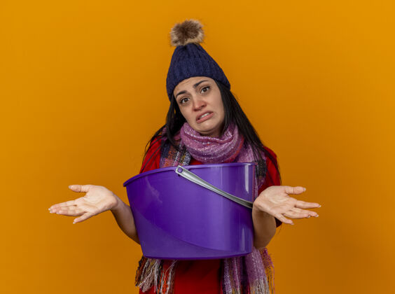 人笨手笨脚的生病的年轻女人戴着冬天的帽子 戴着围巾 手里拿着塑料桶 恶心地看着前面 两手空空的 孤立在橙色的墙上围巾衣服公民