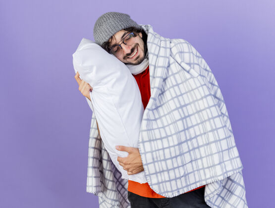人微笑的年轻病夫戴着眼镜 戴着冬天的帽子和围巾 裹着格子呢 抱着枕头 把头放在上面 闭着眼睛 孤立地站在紫色的墙上格子人帽子