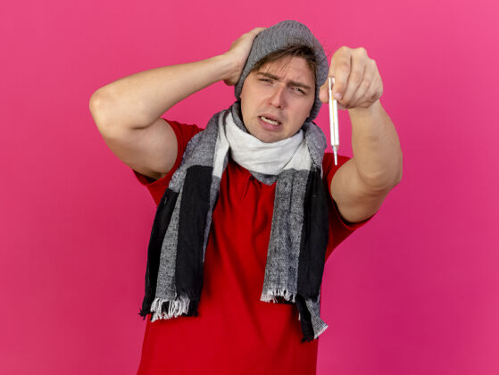 冬天皱眉的年轻英俊的金发病男子戴着冬天的帽子和围巾向前面伸出温度计手放在头上看着前面隔离在粉红色的墙上感觉穿着年轻人