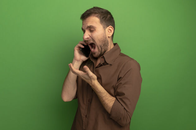 年轻愤怒的年轻人站在侧视图上讲电话 在绿色的墙上显示孤立的空手人人电话