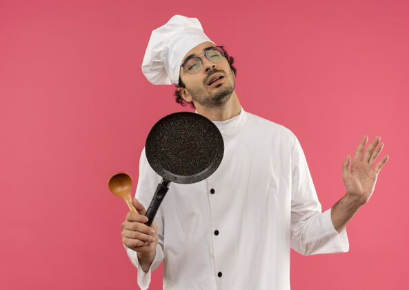 烹饪年轻的男厨师闭着眼睛 穿着厨师制服 戴着眼镜 拿着勺子拿着平底锅 把手上的勺子抹在粉色上背景持有眼睛