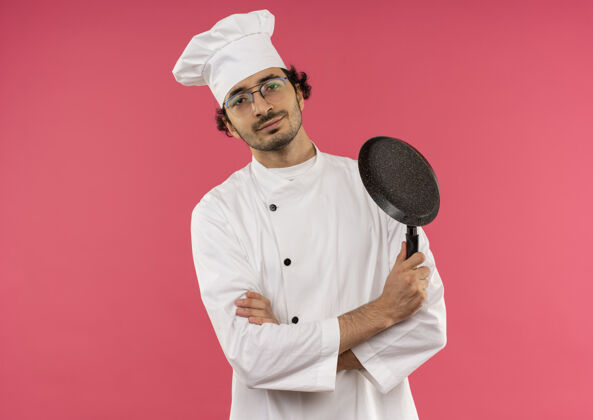 年轻年轻的男厨师穿着厨师制服 戴着眼镜 双手交叉 手里拿着粉红色的煎锅男请眼镜