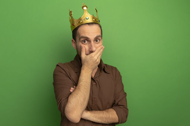 冠惊讶的年轻人戴着皇冠看着前面把手放在嘴上隔离在绿色的墙上人人人