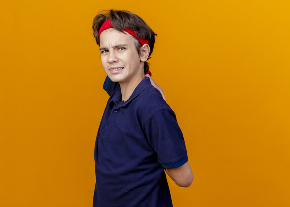 男孩疼痛的年轻英俊的运动男孩戴着头带和戴着牙套的腕带站在侧视图中 手放在背后孤立在橙色的墙上脸穿着年轻
