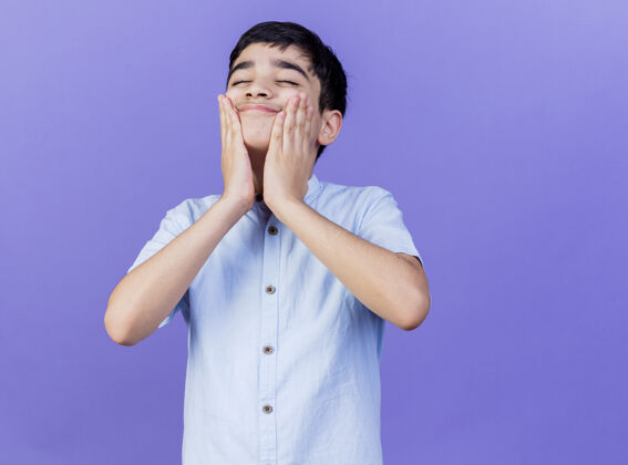 男孩高兴的小男孩手放在脸上闭着眼睛孤立在紫色的墙上手保持人