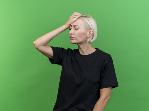 手疼痛的中年金发女人把手放在头上闭着眼睛孤立在绿色的墙上脸市民疼痛