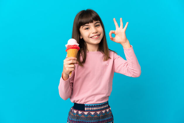 圆锥形小女孩拿着小号冰激凌孤立地站在蓝色的墙上 用手指表示ok表情手认可