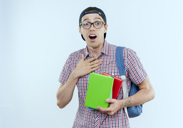 背景惊讶的年轻学生男孩戴着书包 戴着眼镜和帽子拿着书 把手放在白色的胸前胸部帽子学生