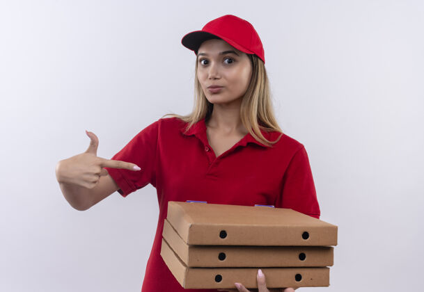 年轻人年轻的送货女孩穿着红色制服 戴着帽子 指着隔离在白色墙上的比萨饼盒帽子送货穿