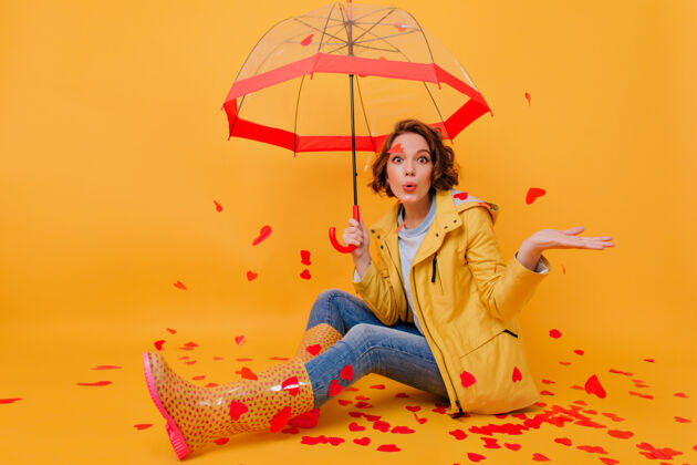 雨穿着胶鞋和蓝色牛仔裤 打着伞的漂亮女孩的室内照片坐在地板上的快乐女士的画像 红色的纸心天气漂亮情绪