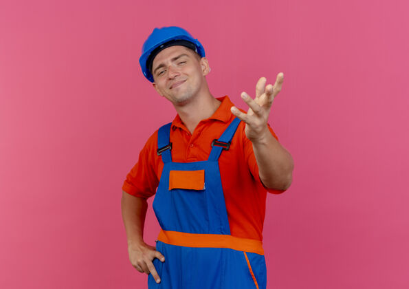 建筑工人年轻的男建筑工人穿着制服 戴着安全帽 手伸向镜头 另一只手放在粉色的臀部安全请男性