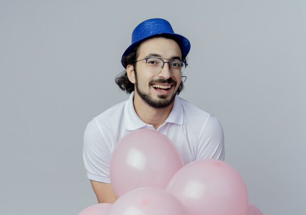 站着笑容可掬的帅哥戴着眼镜和蓝帽子站在白色气球后面孤立男人戴着眼镜