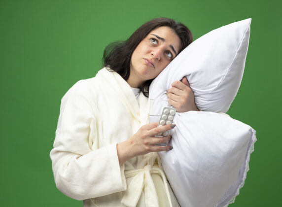 枕头虚弱的年轻白种人病女孩穿着长袍抱着枕头把头放在上面拿着一杯水和一包药片在绿色的背景上孤立地看着虚弱向上玻璃
