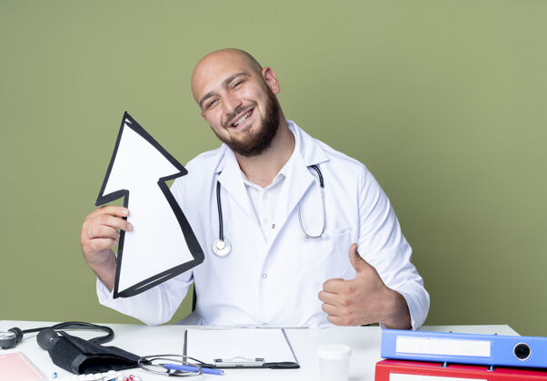 长袍面带微笑的年轻秃头男医生穿着医用长袍和听诊器坐在办公桌前工作坐着方向标记