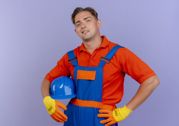紫色高兴的年轻男子建设者穿着制服 戴着手套 戴着安全帽 手放在臀部 紫色隔离建设者手势头盔
