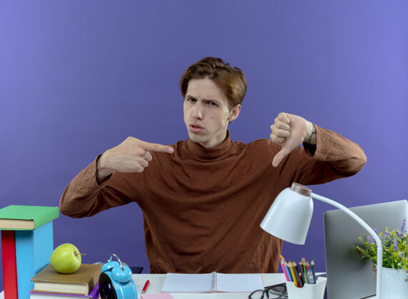 不愉快坐在书桌旁 拿着学习工具的一个没出息的小男孩拇指朝下 指着紫色的拇指学校工具拇指