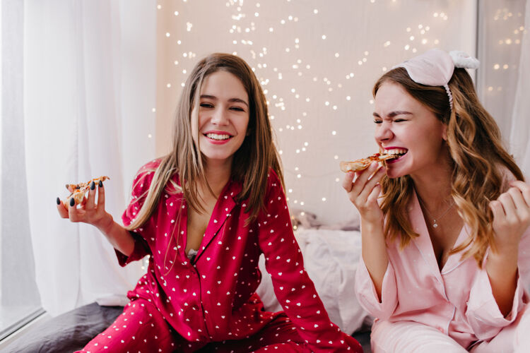 睡衣热情的卷发女孩在吃披萨身着红色睡衣的高加索年轻女子在卧室里拿着快餐摆姿势的室内照片女人舒适年轻