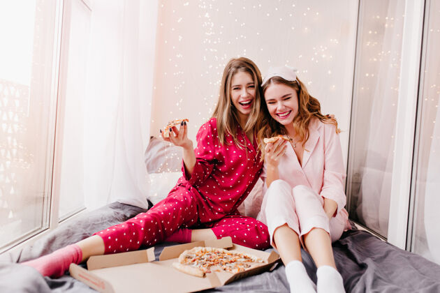 睡衣可爱的女孩穿着时髦的红色睡衣 和姐姐一起享用奶酪披萨微笑着的漂亮女士早上在床上吃快餐情绪眼罩黑发