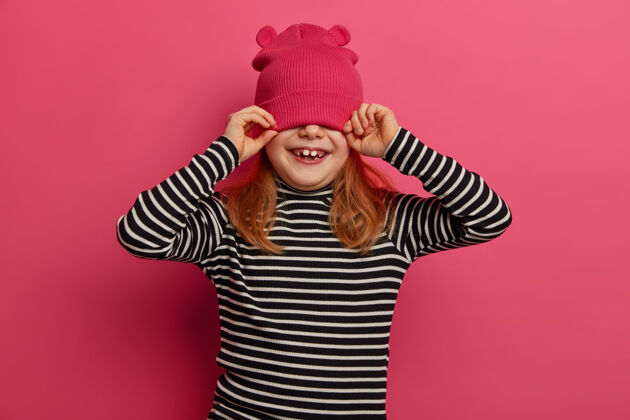 玩四岁女孩穿着条纹套头衫 戴着粉色帽子 玩得开心 蒙着眼睛 喜欢在家庭圈子里度过时光 孤立在粉色的墙上孩子 情感 衣服可爱女孩请