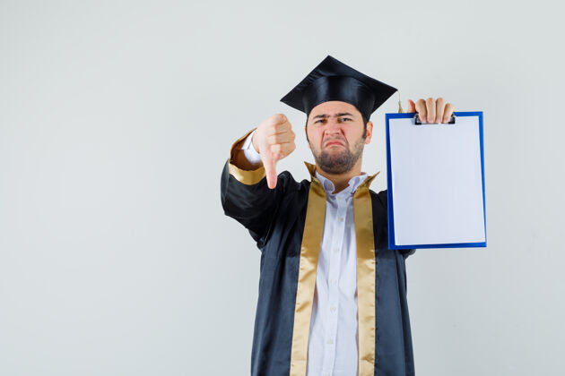 肖像身穿研究生制服的年轻人拇指朝下拿着剪贴板 看上去很不满意 正视图毕业衬衫下来