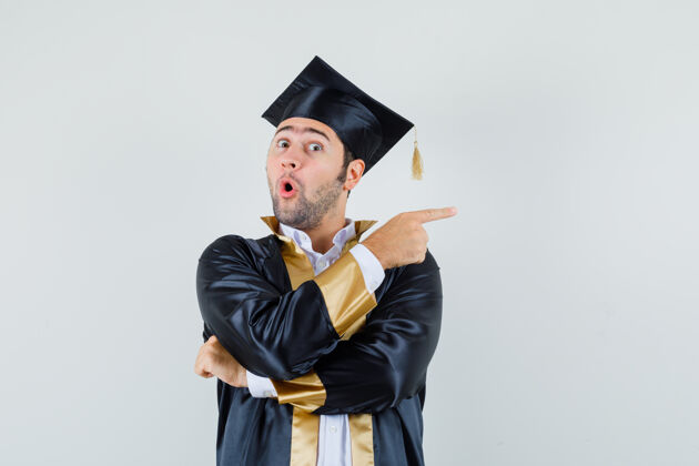 胡须年轻人指着一边穿着毕业制服 看起来很惊讶前视图毕业帅哥欢呼