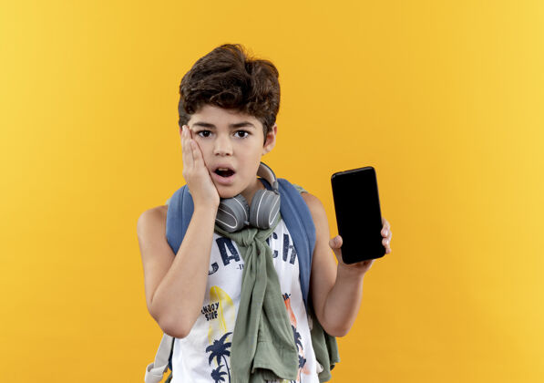 电话惊讶的小男生背着书包 戴着耳机 手里拿着电话 手放在面颊上隔离在黄色上包表情脸颊