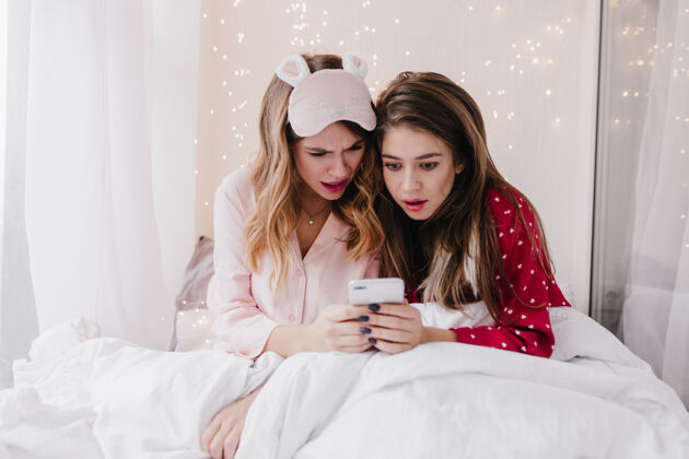 朋友惊讶的欧洲女士坐在床上看着手机屏幕惊讶的穿着睡衣的女孩们在读短信金发舒适女士