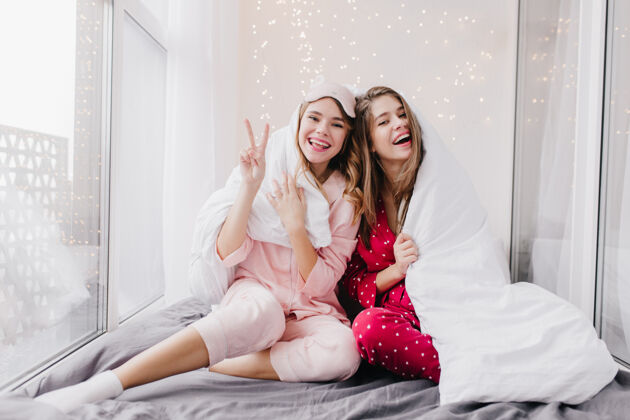 金发穿着睡衣的漂亮卷发女人在卧室里摆姿势表达幸福两个穿着睡衣的欧洲女孩坐在毯子下面睡眠夜晚冷