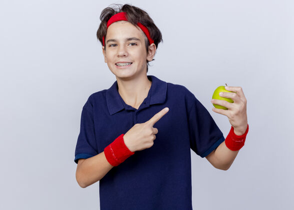 感觉面带微笑的年轻英俊的运动男孩戴着头带和护腕 戴着牙套 看着前面 拿着苹果 指着隔离在白色墙上的苹果市民姿势穿着