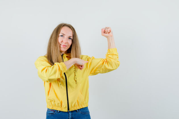肖像穿着黄色棒球服和蓝色牛仔裤的年轻女子展示她的肌肉 指着它 看起来很有力量前视图肌肉可爱夹克