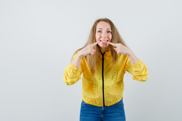 肖像穿着黄色棒球服和蓝色牛仔裤的年轻女子用食指指着嘴巴 看上去很乐观 正面视图成人女性年轻