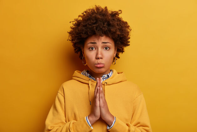 悲伤请允许我哀求的非洲裔美国妇女请求允许 牵手祈祷 说原谅我 对着黄色的墙壁摆姿势 穿着运动衫乞求并说原谅我恳求不高兴祈祷