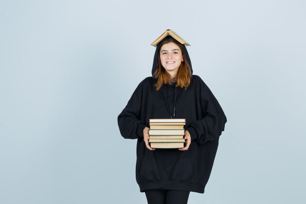 头部年轻的女士举着书在头顶上作为屋顶 拿着书在她面前的超大连帽衫 裤子和看起来很有趣前视图持有成人漂亮