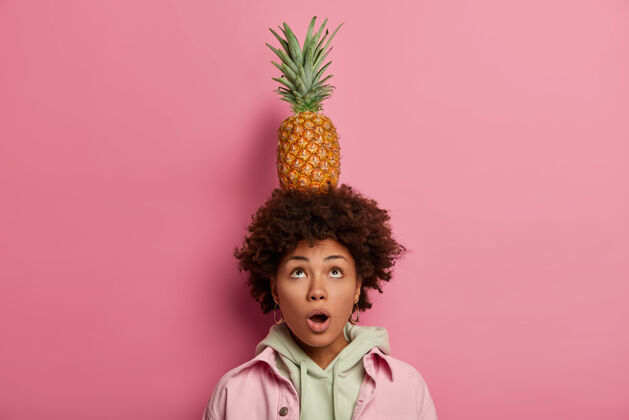 女人惊讶的卷发非洲裔美国妇女抬头望去 张着嘴 头上顶着菠萝 想知道她怎么能做到 穿着连帽衫和夹克 对着粉色粉彩墙摆姿势热带时尚菠萝
